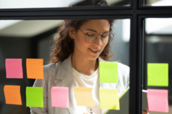 A woman looks over her organized sticky notes on a window as part of her project management A woman looks over her organized sticky notes on a window as part of her project management