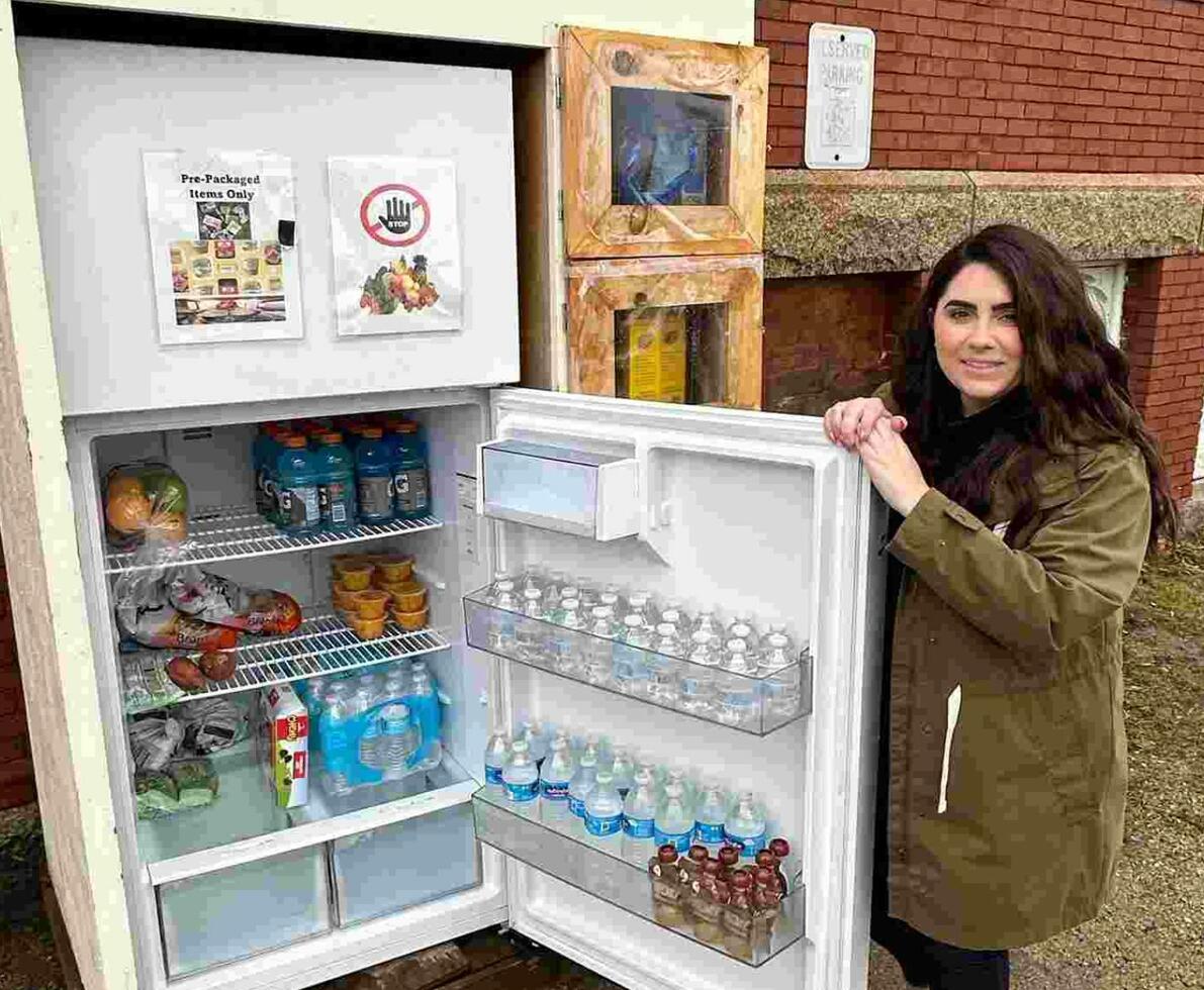 A woman posing in front of an open community refrigerator outside of a brick building