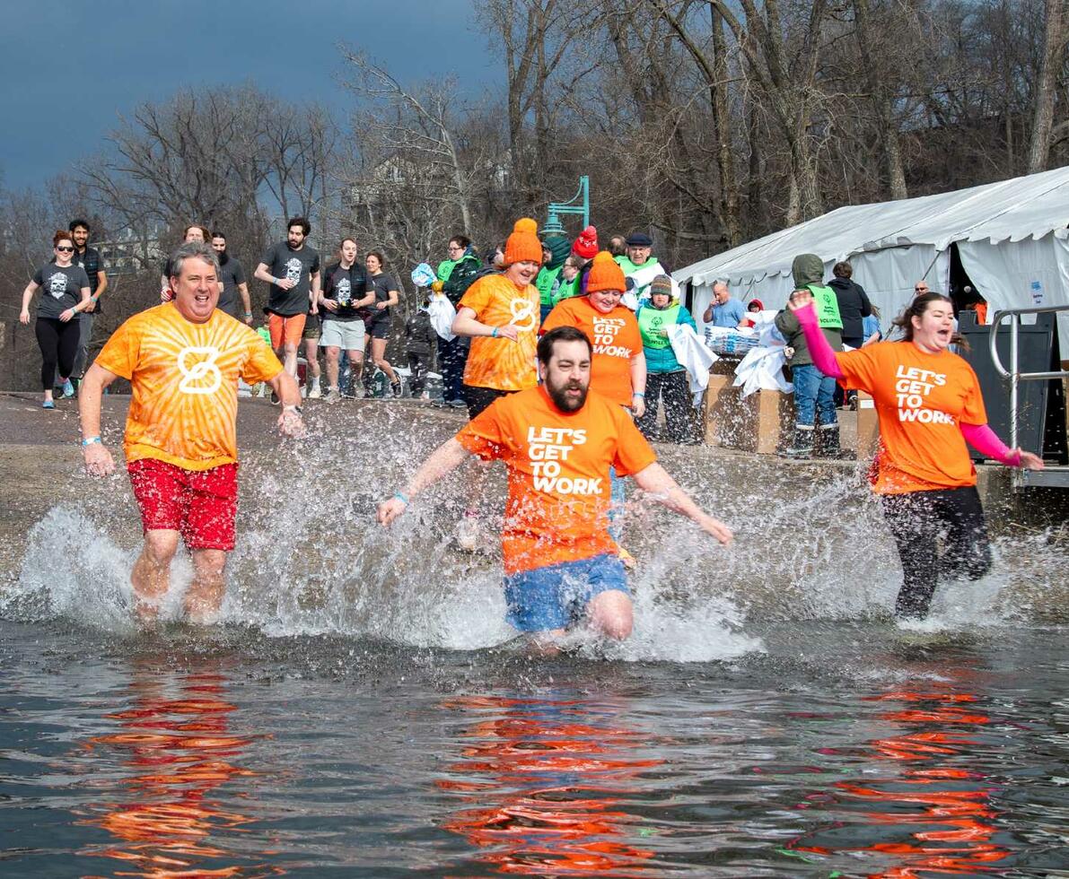 Three people in orange t-shirts running and splashing into a lake. White tents and more people are in the background.