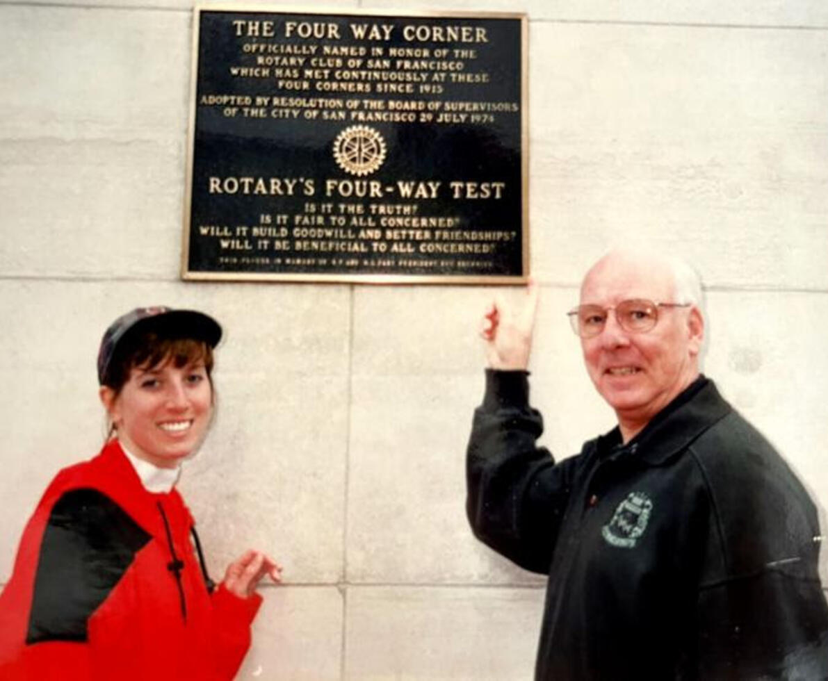 Two people standing up smiling at the camera. The woman wears a red jacket. The man wears a black jacket and is pointing upward to a plaque on a white wall.
