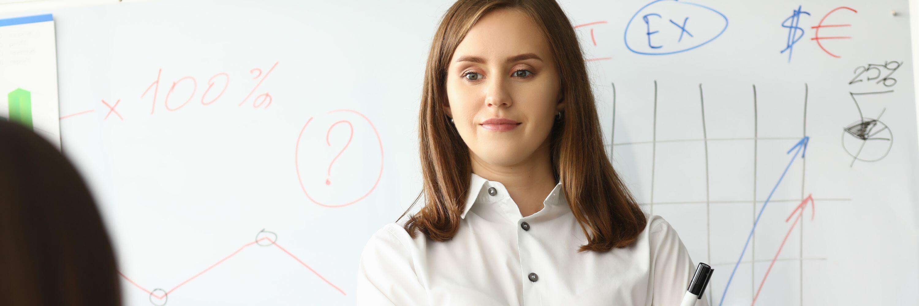A woman in white top with brown hair standing in front of a whiteboard with notes on it