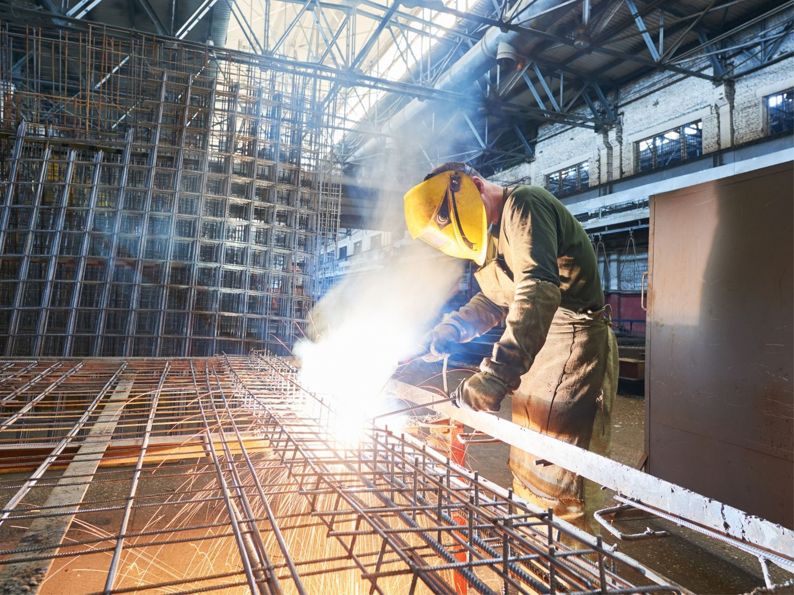 Welder wearing a yellow protective mask and using a heat tool to create a metal grid in a warehouse setting.