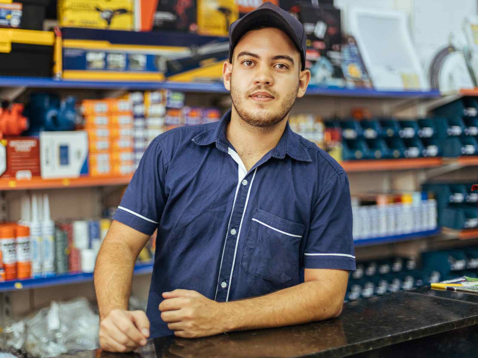  A salesperson wearing a blue shirt and black cap is leaning on a counter in a hardware store.