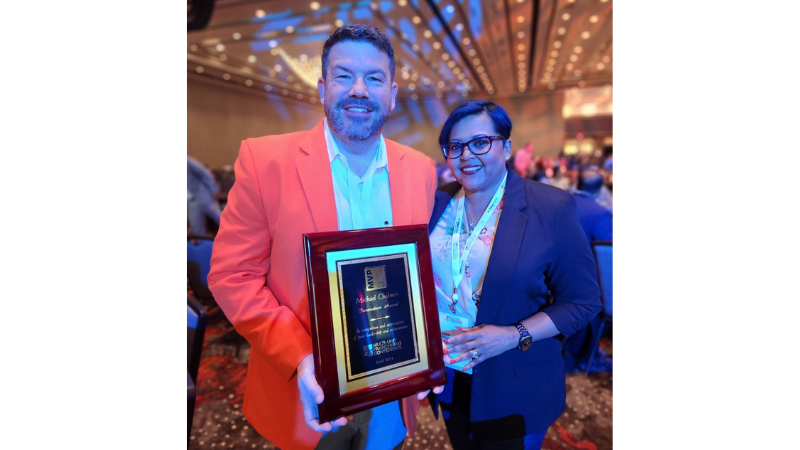 Michael Chalmers, in orange sport coat, holding a plaque, next to Kathy George, in navy blazer, both smiling at the camera