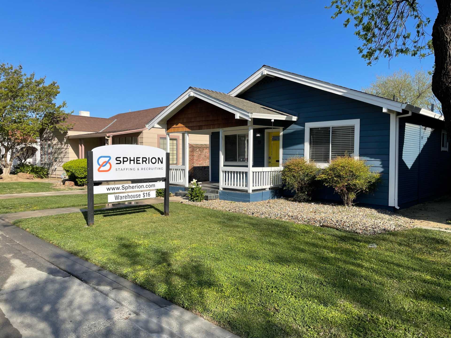 A wide angle view of a blue A-frame house with a green lawn in front. A white sign in the yard reads Spherion Staffing & Recruiting