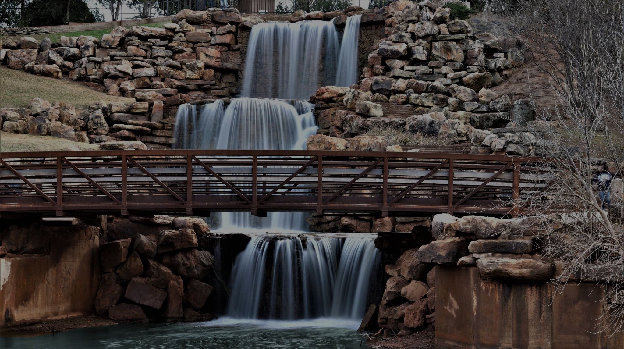Photo of a three-tier waterfall over rocks with a pedestrian bridge in front.