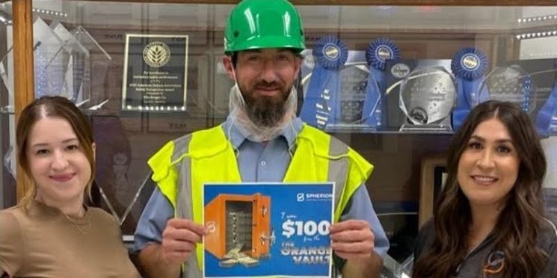 A man in a green hard hat and a yellow safety vest holding a $100 certificate in the middle flanked by two women in office attire.