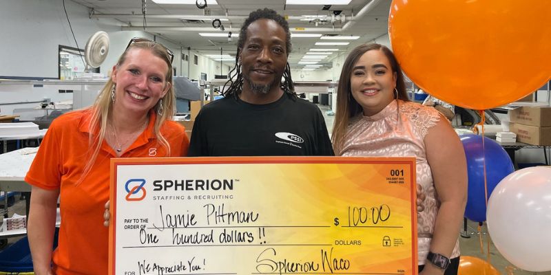 An African-American man holds a giant orange check for $100 flanked by two women in office war in a factory setting