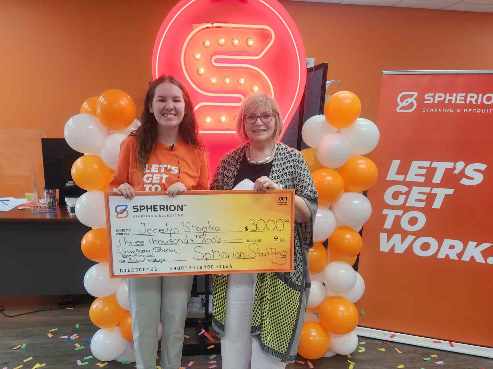 A girl and a woman smiling while holding a giant scholarship check, standing in front of an orange and white balloon installation and a neon S sign.