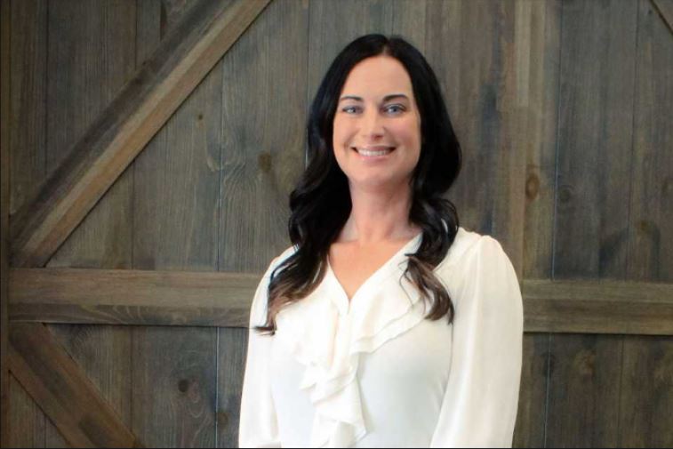 A close-up of a woman smiling in a white shirt with a wooden wall background