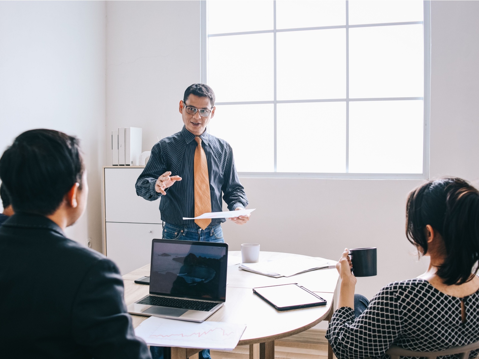 A male CEO standing in a white office speaks to another male and a woman seated in the foreground