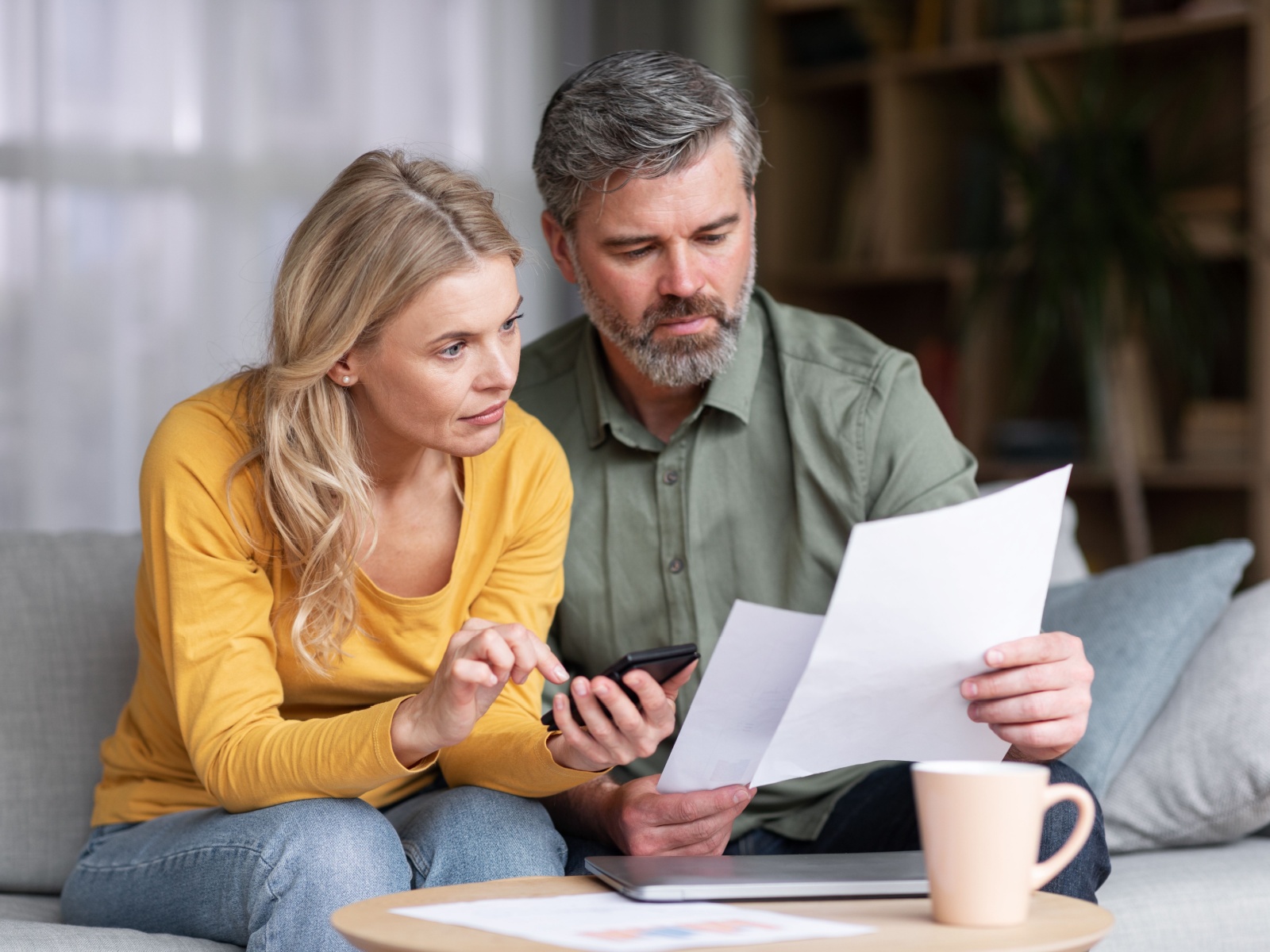 Married Middle Aged Couple Planning Budget Together, Reading Papers And Calculating Spends While Sitting On Couch In Living Room