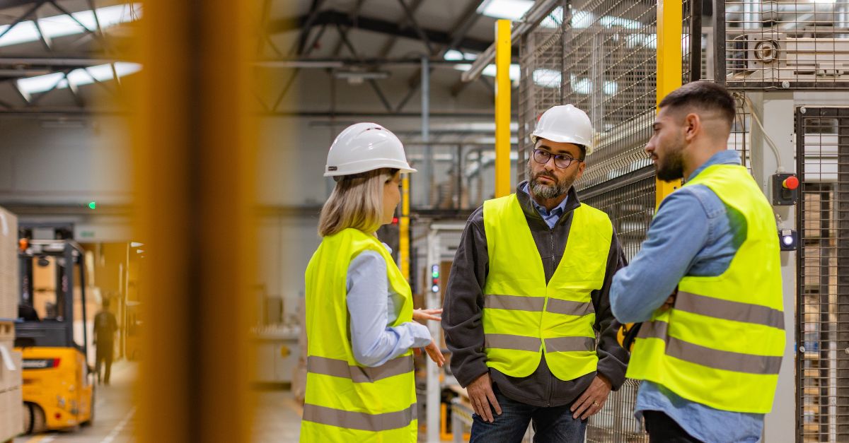 Three workers in safety vests and hard hats talking in a warehouse