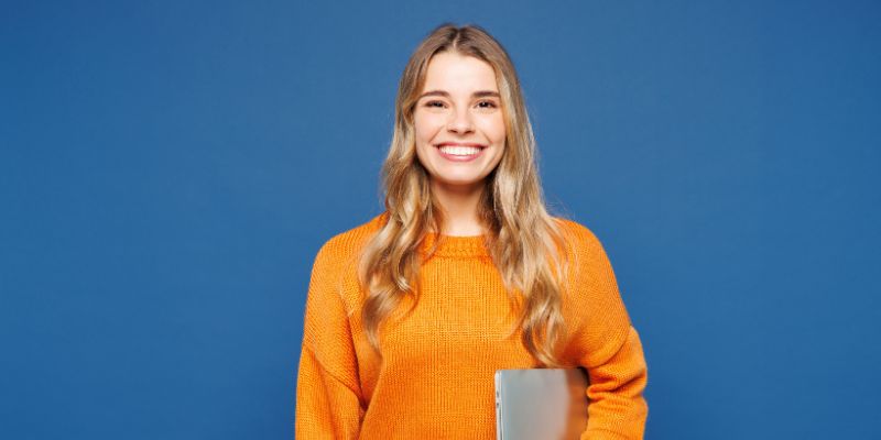 Young woman in an orange sweater smiling and holding a laptop