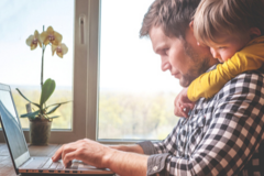 Dad typing on a laptop with his son hanging around his shoulders