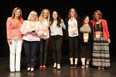 A group of seven women smiling at the camera and accepting awards on stage