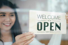 Asian American woman holding a sign that reads Yes We're Open in a storefront window