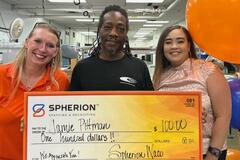 An African-American man holds a giant orange check for $100 flanked by two women in office war in a factory setting