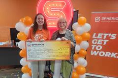 A girl and a woman smiling while holding a giant scholarship check, standing in front of an orange and white balloon installation and a neon S sign.