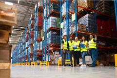 Warehouse staff meeting, workers standing with managers in front of floor-to-ceiling shelves of goods