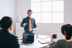 A male CEO standing in a white office speaks to another male and a woman seated in the foreground
