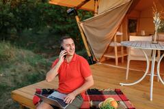 Happy businessman freelance using laptop while talking by phone on cozy glamping tent in a summer day.