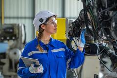 A woman in a blue uniform and white hard hat inspecting a factory machine