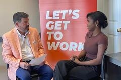 A man interviewing a young woman with an orange "Let's get to work" sign in the background