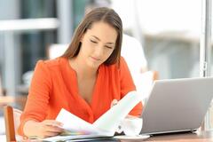 A woman in an orange shirt looking at paperwork and a laptop