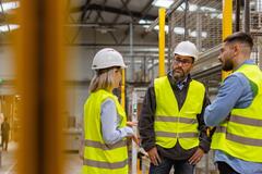 Three workers in safety vests and hard hats talking in a warehouse