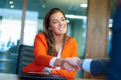A woman in an orange jacket smiling and shaking hands with someone off camera