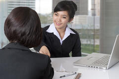 An Asian woman smiles and speaks to a female job candidate during an interview in an office.