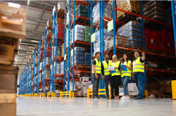 Warehouse staff meeting, workers standing with managers in front of floor-to-ceiling shelves of goods