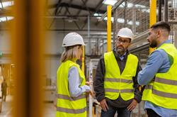 Three workers in safety vests and hard hats talking in a warehouse