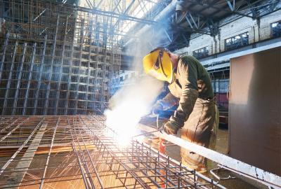 Welder wearing a yellow protective mask and using a heat tool to create a metal grid in a warehouse setting.