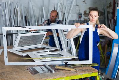 A woman, foreground, and a man, background, assemble white metal frames at work.