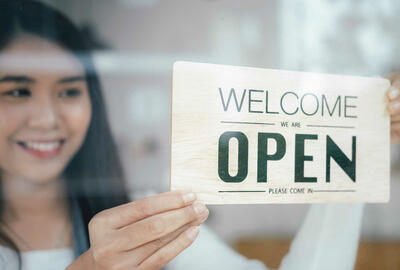 Asian American woman holding a sign that reads Yes We're Open in a storefront window