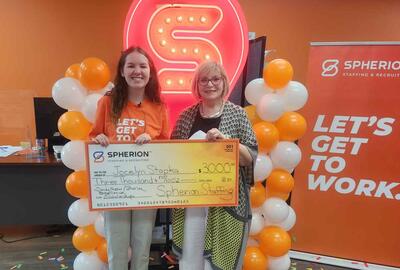 A girl and a woman smiling while holding a giant scholarship check, standing in front of an orange and white balloon installation and a neon S sign.