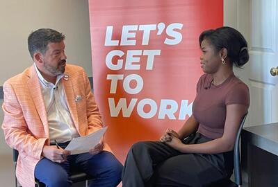 A man interviewing a young woman with an orange "Let's get to work" sign in the background