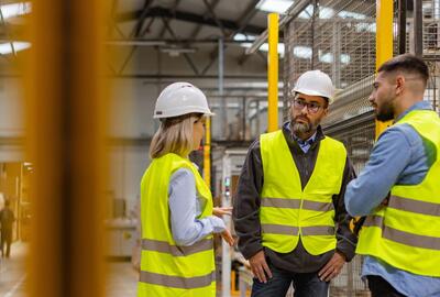 Three workers in safety vests and hard hats talking in a warehouse