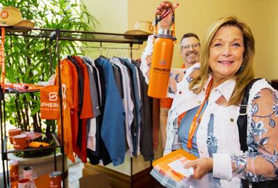 Photo of a woman smiling and holding up an orange water bottle in front of a rack of clothing