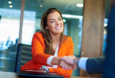A woman in an orange jacket smiling and shaking hands with someone off camera