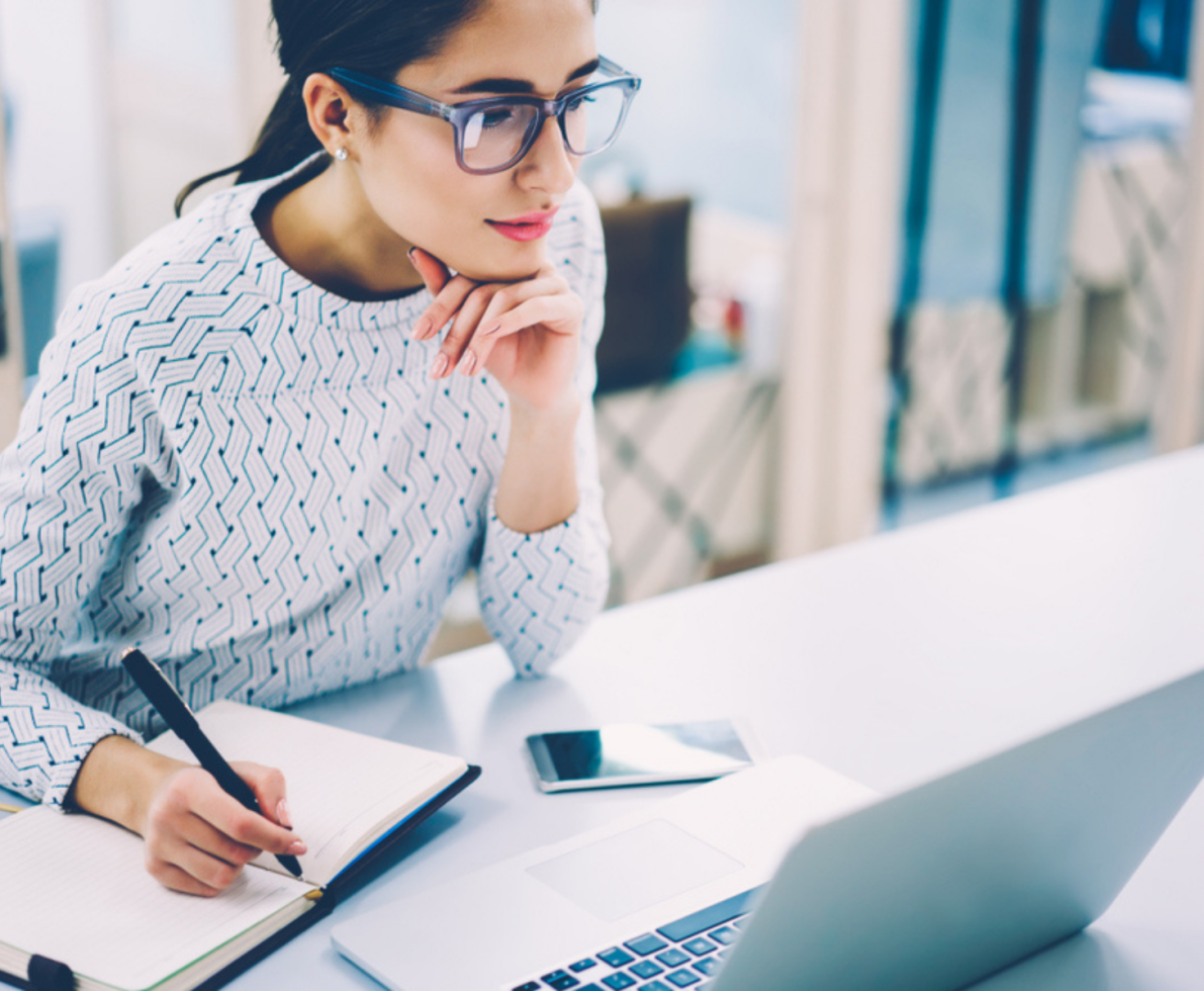 A woman in a white short and glasses takes notes while looking at a laptop screen