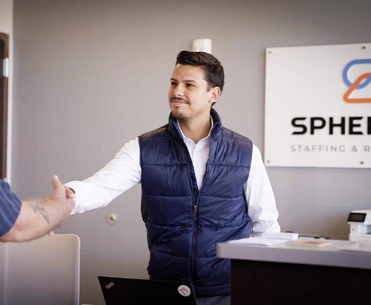 Man in navy vest shaking hands with a person off camera. Sign in background reads Spherion Staffing & Recruiting