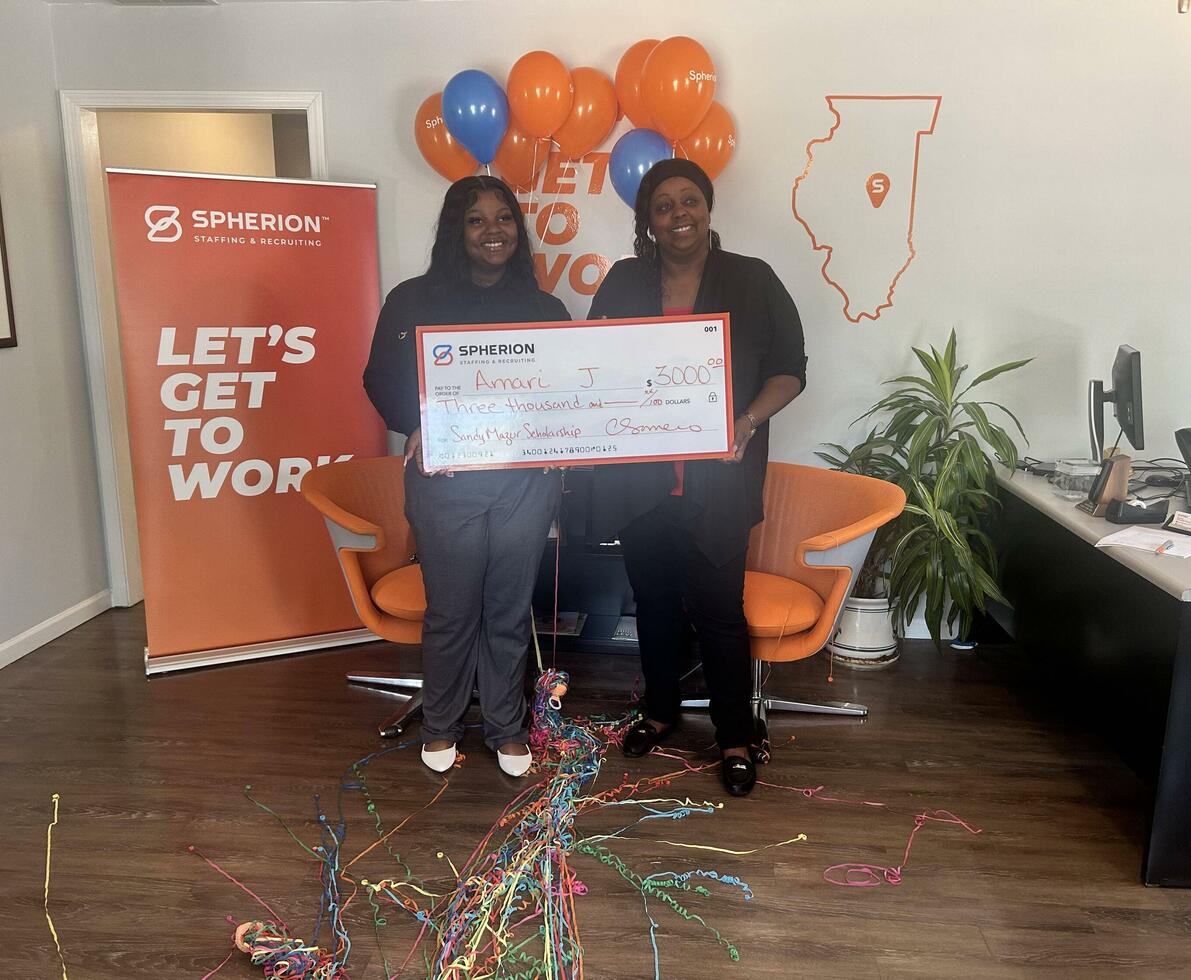 Two women holding a giant scholarship check in front of an orange banner that reads Let's Get to Work