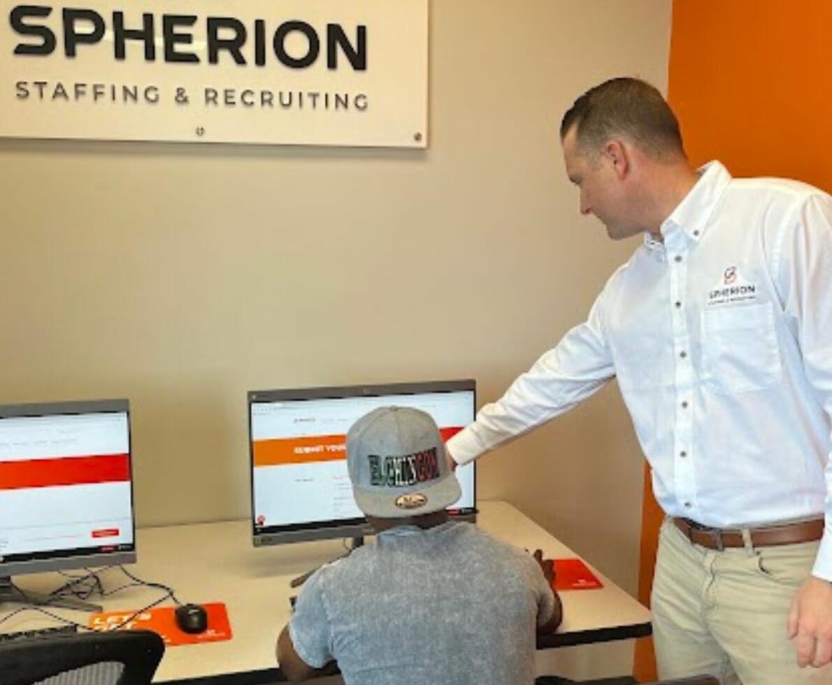 A man seated at a computer is shown how to apply for a job by another man standing to his right.