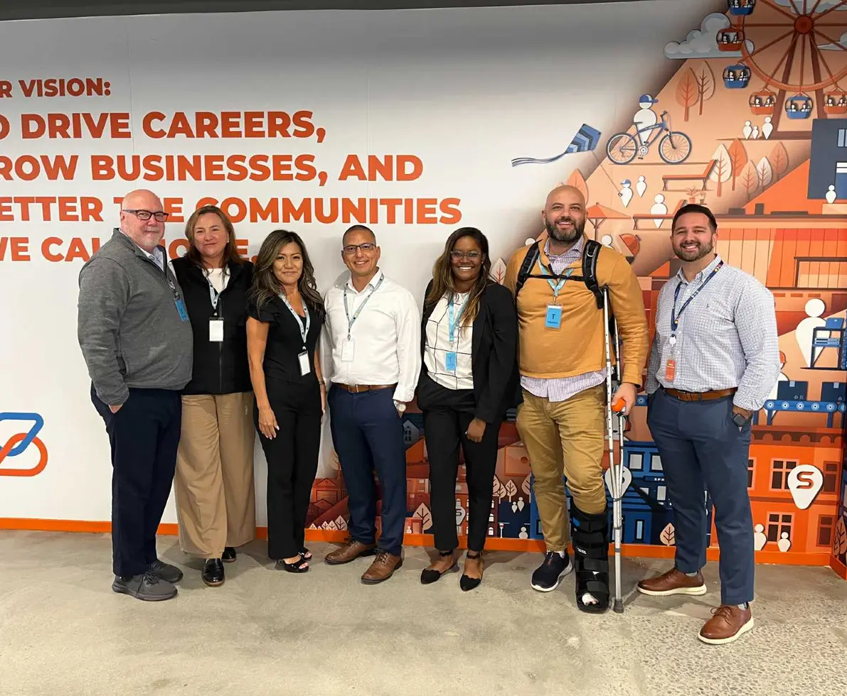 Photo of a group of smiling people standing in front of a white, orange and blue wall mnural