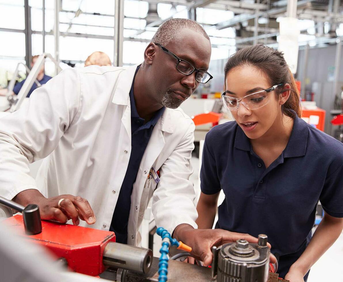 Black man in a lab coat next to a Hispanic woman in a blue shirt inspecting a piece of equipment