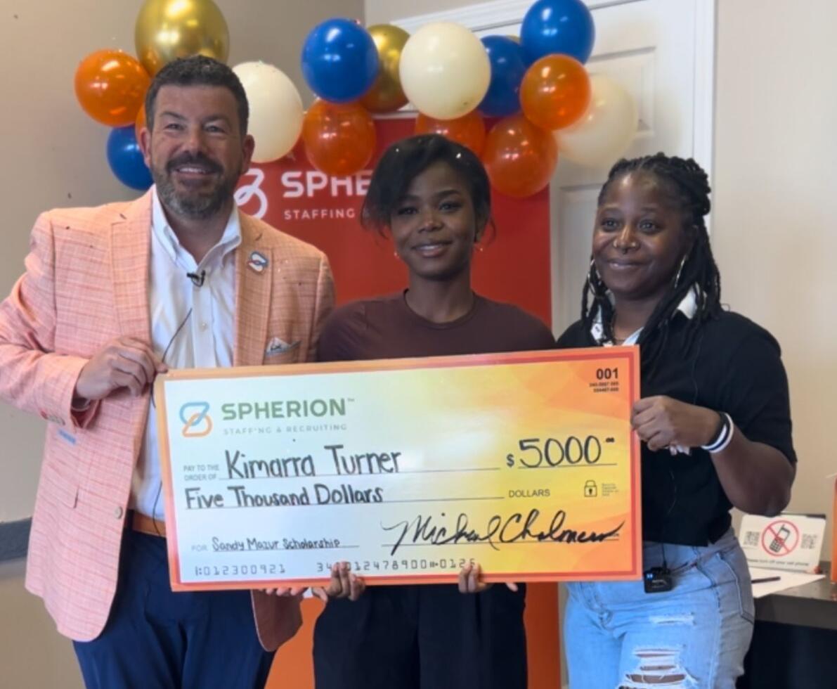 Three people smiling and standing behind a giant orange check in an office setting