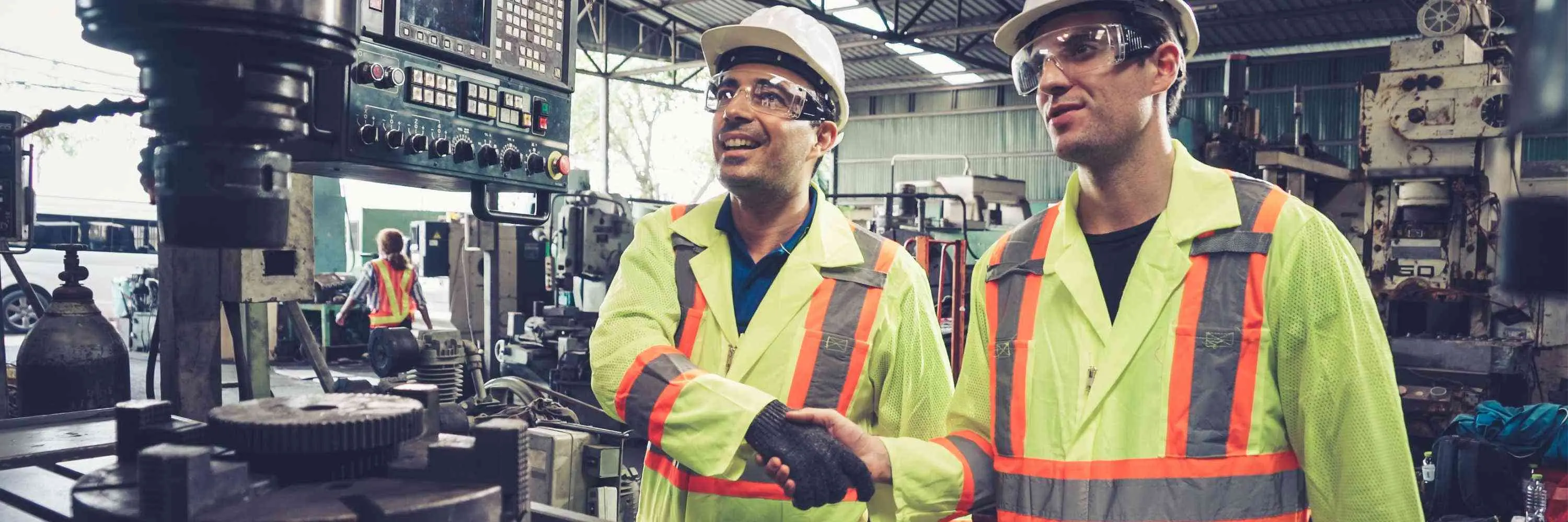 Two machinists in yellow safety vests shaking hands and smiling at the success of a working machine